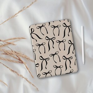 Könnte beinhalten: Eine beige iPad-Hülle mit einem schwarzen Schleifenmuster. Die Hülle liegt auf einer weißen Oberfläche.