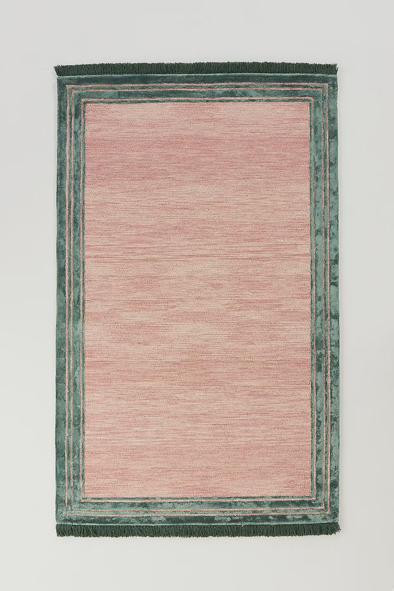May include: Rectangular rug with a pink center and a teal green border. The rug has a textured appearance, with the pink area showing horizontal lines. The border is a darker teal green, with a fringe along the edge.