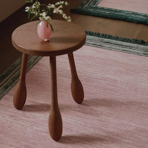 May include: A small, round wooden side table with three legs stands on a pink rug. A small pink vase with white flowers sits on the table. The rug has a green border.