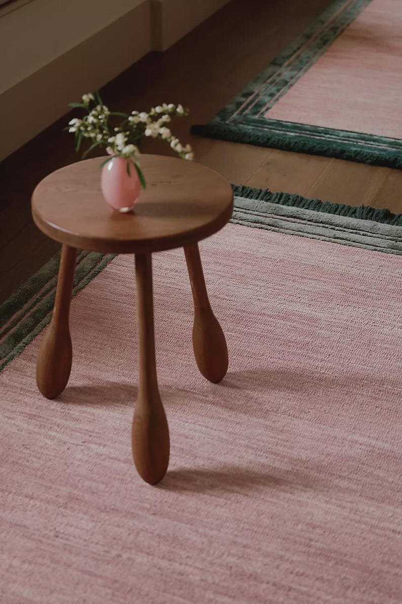 May include: A small, round wooden side table with three legs stands on a pink rug. A small pink vase with white flowers sits on the table. The rug has a green border.