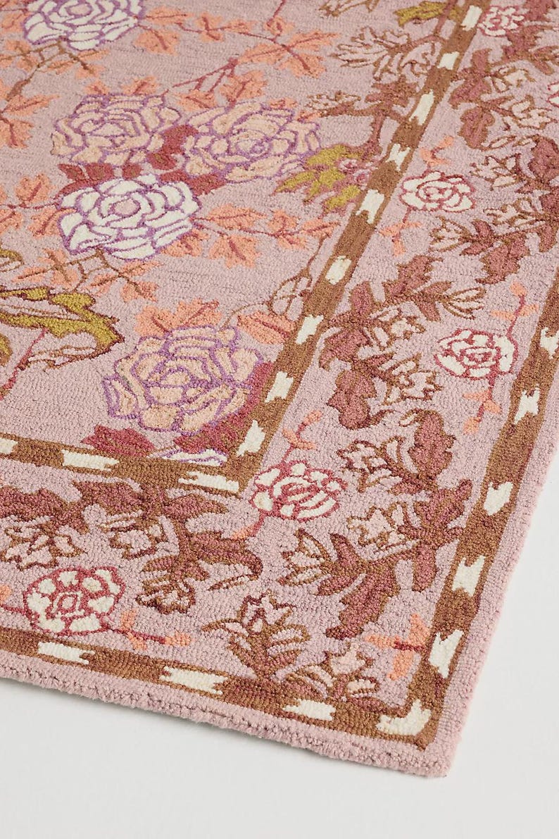 May include: A rectangular rug with a floral pattern in shades of pink, brown, and white. The rug features a repeating design of roses and leaves, with a border of brown and white accents. The rug is made of a soft, textured material.