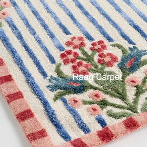 May include: A rectangular rug with a cream background, blue stripes, and floral designs in red, pink, and green. The border is pink and red. The text "Raag Carpet" is visible.