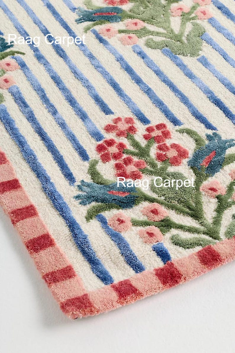 May include: A rectangular rug with a cream background, blue stripes, and floral designs in red, pink, and green. The border is pink and red. The text "Raag Carpet" is visible.