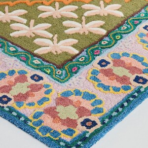 May include: A colorful, patterned rug with a botanical theme. The rug features a green background with dragonfly and lily pad designs. The border has floral patterns in pink, blue, and yellow, with a blue trim. The rug is made of a soft, textured material.