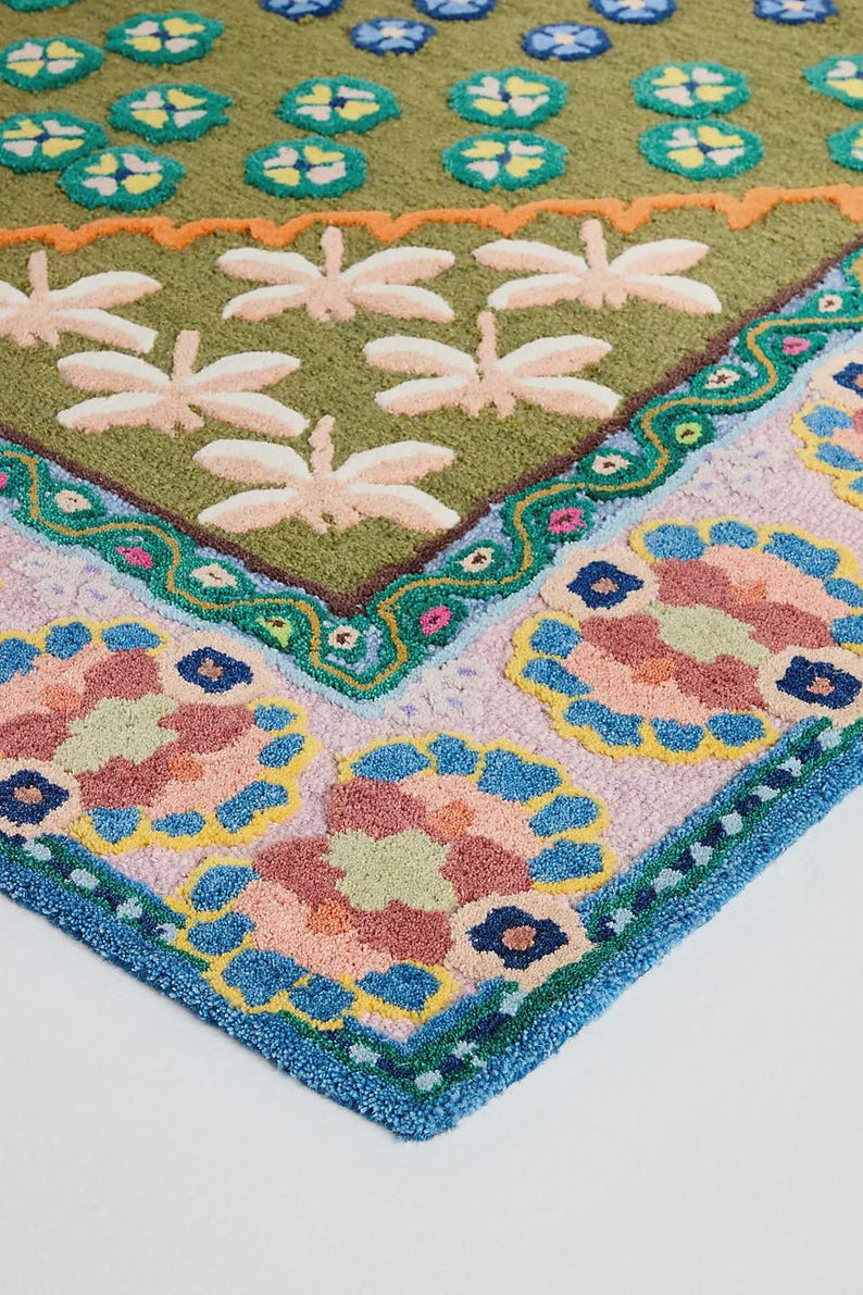 May include: A colorful, patterned rug with a botanical theme. The rug features a green background with dragonfly and lily pad designs. The border has floral patterns in pink, blue, and yellow, with a blue trim. The rug is made of a soft, textured material.