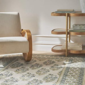 May include: A cream-colored rug with a floral pattern in shades of blue and green. The rug is placed in a room with a cream-colored chair and a gold side table. The rug has a patterned border.