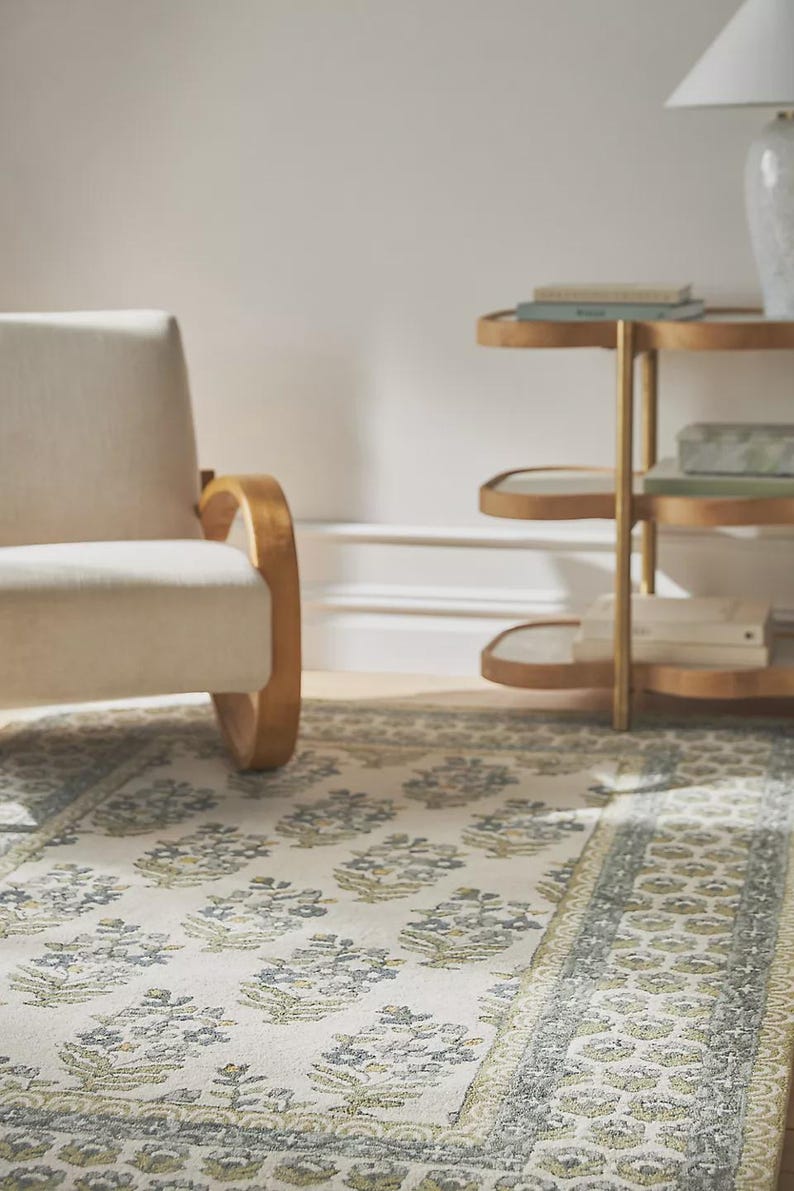 May include: A cream-colored rug with a floral pattern in shades of blue and green. The rug is placed in a room with a cream-colored chair and a gold side table. The rug has a patterned border.