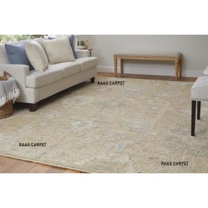 May include: A beige area rug with a faded floral pattern dominates the room. The rug is placed in front of a white sofa with blue and cream-colored pillows. A wooden bench and a chair are also in the room. The text "RAAG CARPET" is printed on the rug.
