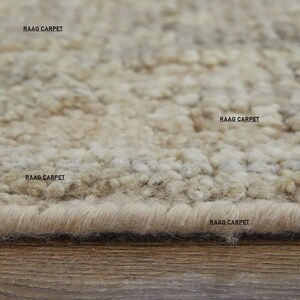 May include: Close-up of a beige area rug with a textured, looped pile. The rug has a neutral color palette with variations of beige and tan. The text "RAAG CARPET" is printed on the rug in several places.