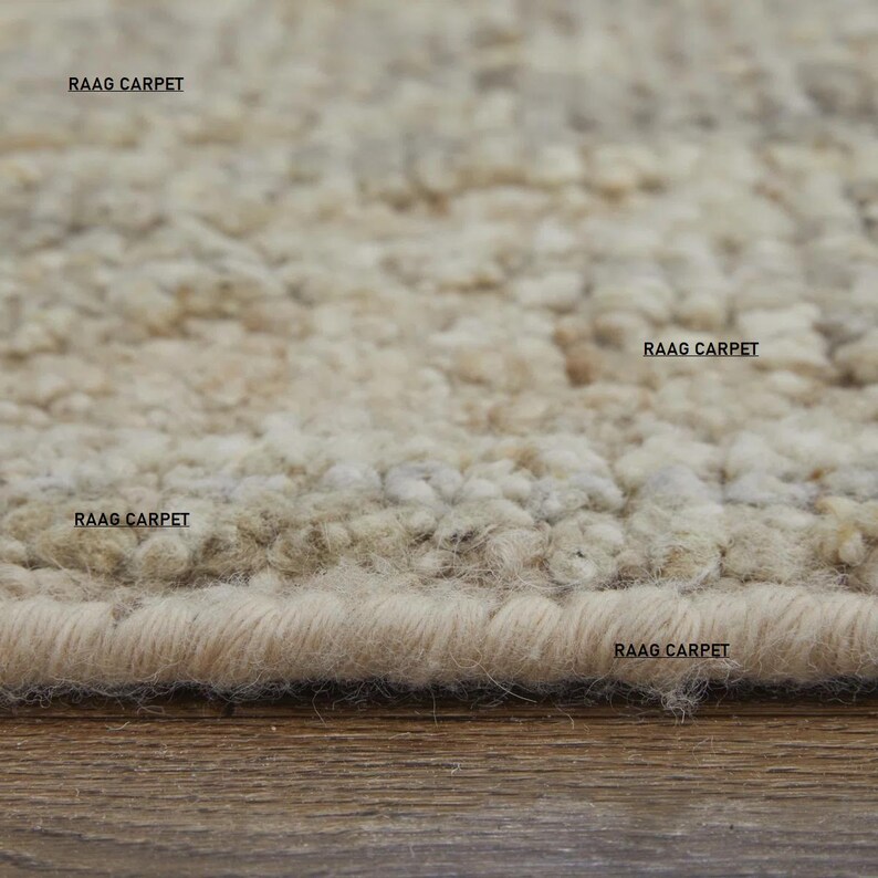 May include: Close-up of a beige area rug with a textured, looped pile. The rug has a neutral color palette with variations of beige and tan. The text "RAAG CARPET" is printed on the rug in several places.