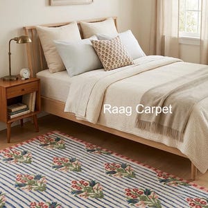 May include: A bedroom scene featuring a striped rug with floral accents in red, green, and blue. The rug is placed in front of a wooden bed with cream-colored bedding and pillows. A wooden nightstand with a lamp and books is visible.