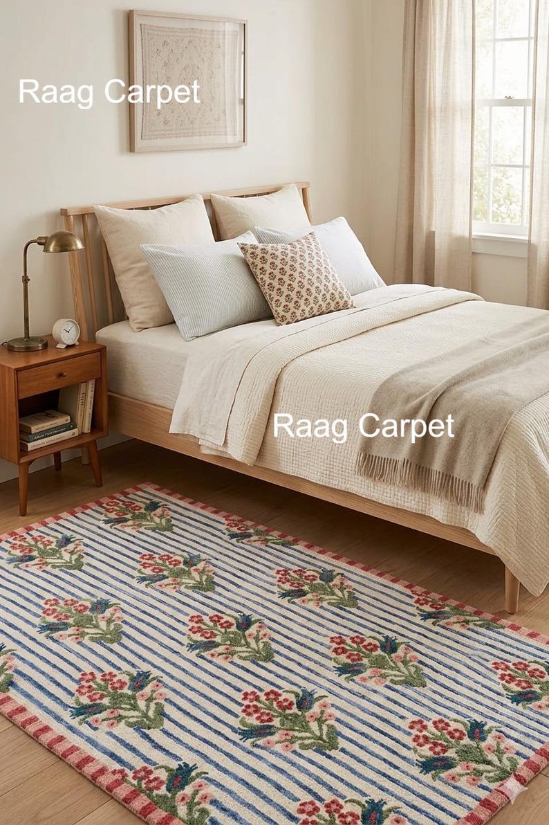 May include: A bedroom scene featuring a striped rug with floral accents in red, green, and blue. The rug is placed in front of a wooden bed with cream-colored bedding and pillows. A wooden nightstand with a lamp and books is visible.