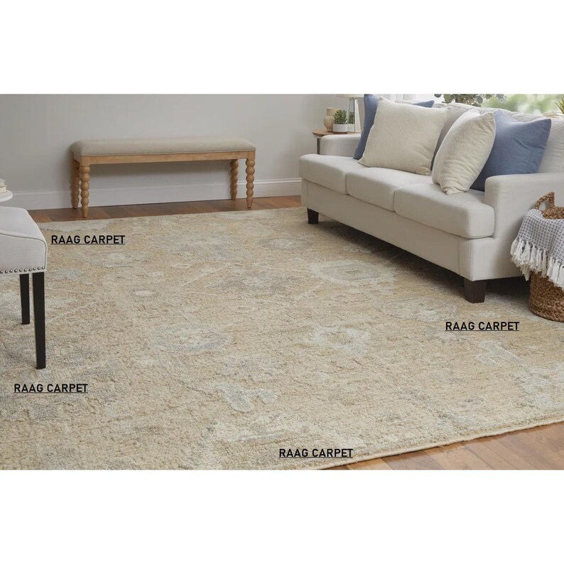 May include: A beige area rug with a faded floral pattern. The rug is in a living room setting, with a sofa, bench, and side table visible. The text "RAAG CARPET" is printed on the rug.