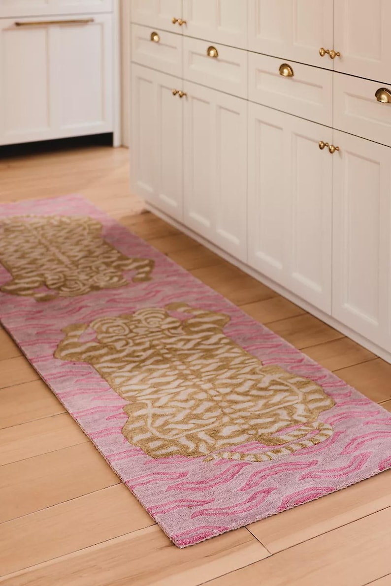 May include: A pink and gold runner rug with a tiger print design. The rug has a pink background with a wavy pattern and two large gold tiger shapes. The rug is placed on a wooden floor.