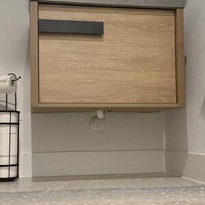 May include: A light wood-grain bathroom vanity with a dark gray countertop and a black handle. The vanity is mounted on the wall above a white baseboard. A black metal toilet paper holder is on the left.