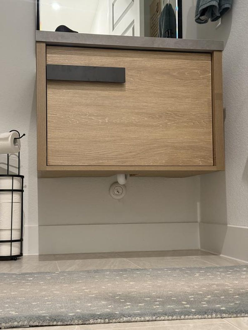 May include: A light wood-grain bathroom vanity with a dark gray countertop and a black handle. The vanity is mounted on the wall above a white baseboard. A black metal toilet paper holder is on the left.
