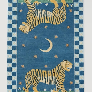 Op de afbeelding: Een rechthoekig tapijt met een blauwe achtergrond met twee tijgers met gele en zwarte strepen. Het tapijt heeft een rand met een geblokt patroon in blauw en wit. Het ontwerp bevat sterren en een maan.