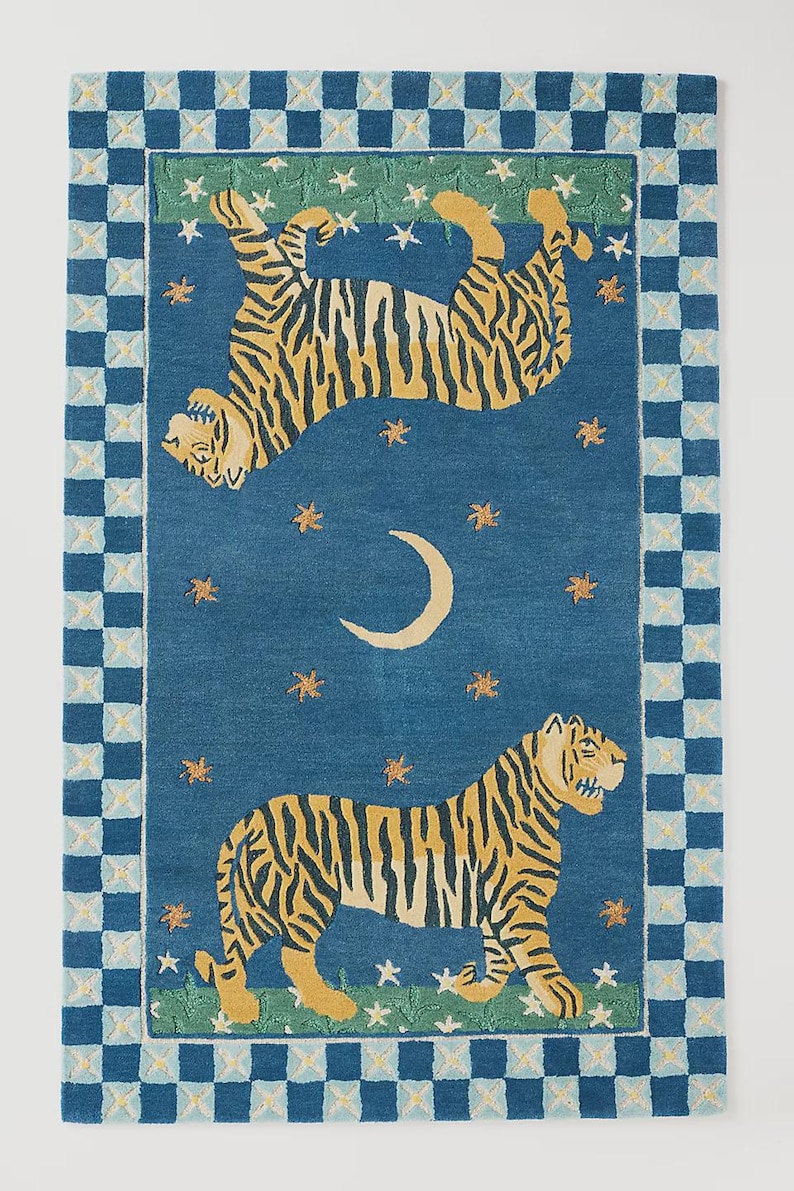 Op de afbeelding: Een rechthoekig tapijt met een blauwe achtergrond met twee tijgers met gele en zwarte strepen. Het tapijt heeft een rand met een geblokt patroon in blauw en wit. Het ontwerp bevat sterren en een maan.