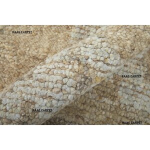 May include: Close-up of a beige and tan textured carpet with a rolled edge. The carpet has a nubby, woven appearance. The text "RAAG CARPET" is visible in multiple places on the carpet.