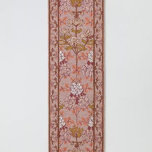 May include: A rectangular rug with a floral design. The rug features a light mauve background with a repeating pattern of white and burgundy roses, and stylized green and gold floral elements. The border is a darker brown with floral accents.
