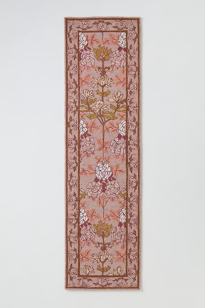 May include: A rectangular rug with a floral design. The rug features a light mauve background with a repeating pattern of white and burgundy roses, and stylized green and gold floral elements. The border is a darker brown with floral accents.
