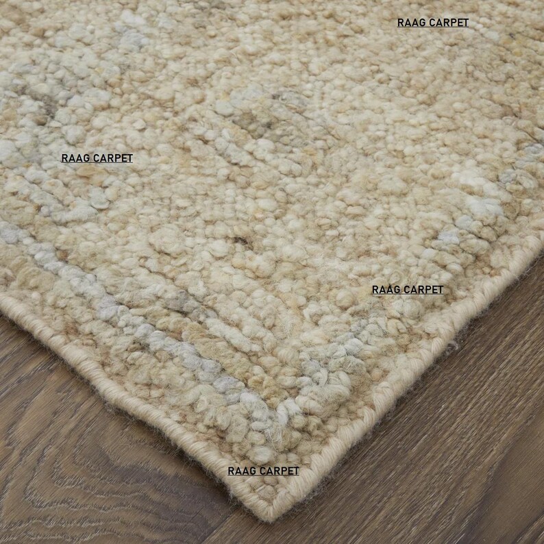 May include: Close-up of a beige textured rug with a geometric pattern. The rug has a thick pile and a neutral color palette, with the text "RAAG CARPET" visible in several places.