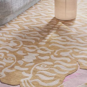 May include: A beige and white patterned area rug with a pink border. The rug features a stylized design with flowing, organic shapes. A gray sofa and white table are partially visible in the background.