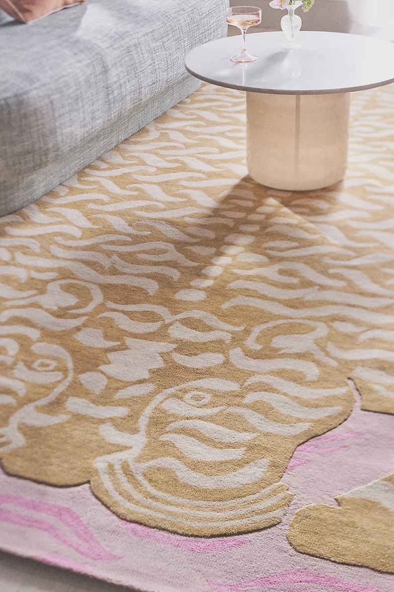 May include: A beige and white patterned area rug with a pink border. The rug features a stylized design with flowing, organic shapes. A gray sofa and white table are partially visible in the background.