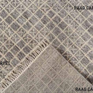 May include: A close-up of a gray and cream-colored rug with a geometric diamond pattern. The rug has a textured surface and a fringed edge. The text "RAAG CARPET" is visible in the image.