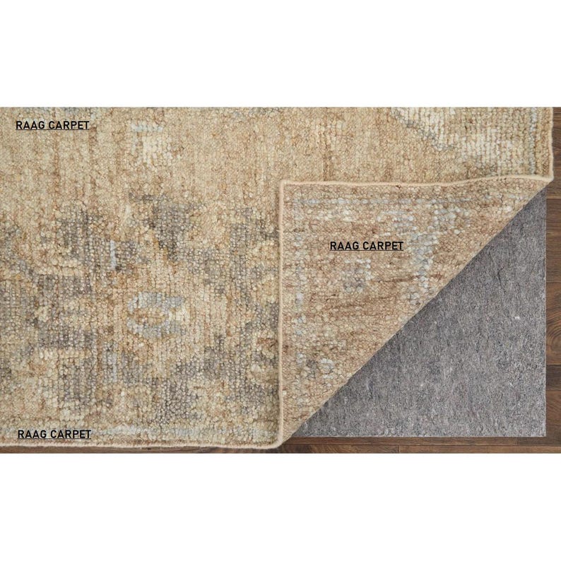 May include: A rectangular beige rug with a distressed pattern, partially rolled back to reveal a gray rug pad. The rug has a faded, vintage look with hints of gray and brown. The text "RAAG CARPET" is visible on the rug.