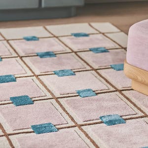 Puede incluir: Una alfombra rectangular con un patr&oacute;n geom&eacute;trico. La alfombra presenta una cuadr&iacute;cula de cuadrados en rosa claro y beige, con cuadrados de acento azul turquesa. L&iacute;neas marrones definen la cuadr&iacute;cula. Una cubierta decorativa para el suelo.