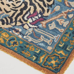 Puede incluir: Secci&oacute;n de una alfombra azul y beige con dise&ntilde;o de tigre. La alfombra presenta un borde verde azulado con patrones geom&eacute;tricos naranjas y amarillos. Las rayas del tigre son negras y la alfombra tiene una apariencia texturizada.