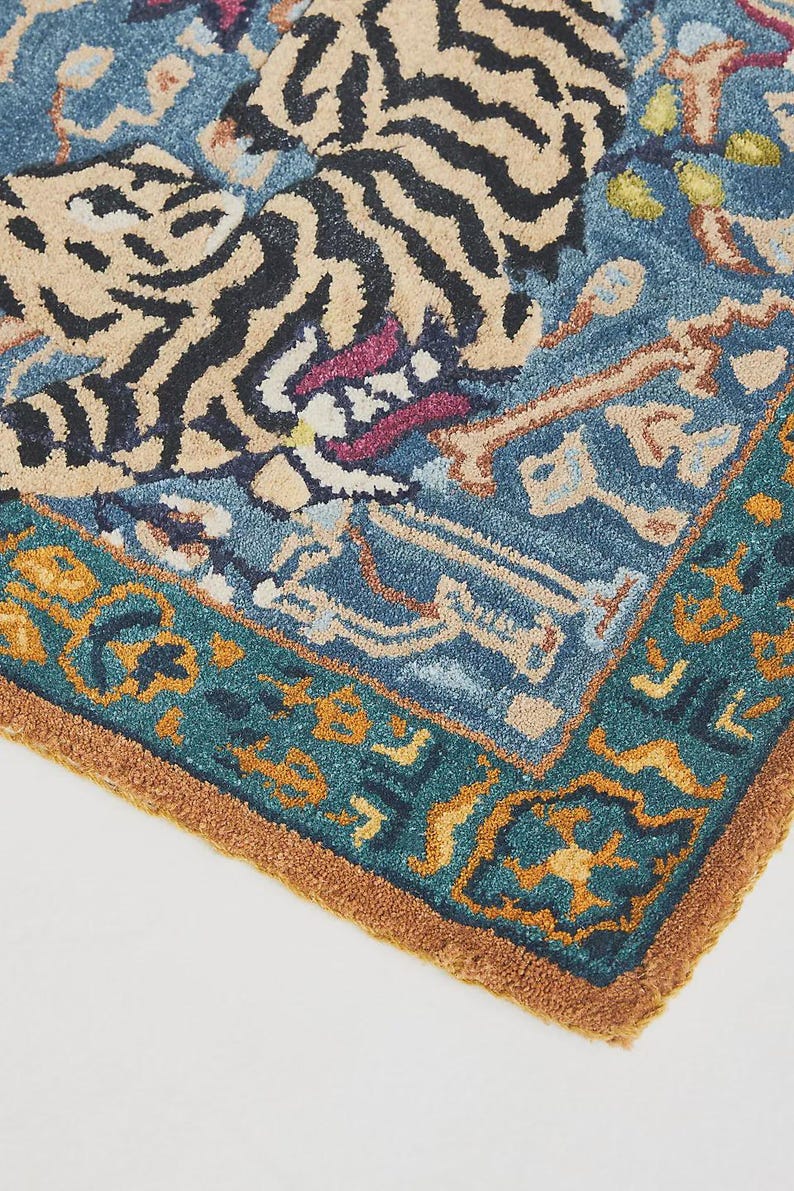 Puede incluir: Secci&oacute;n de una alfombra azul y beige con dise&ntilde;o de tigre. La alfombra presenta un borde verde azulado con patrones geom&eacute;tricos naranjas y amarillos. Las rayas del tigre son negras y la alfombra tiene una apariencia texturizada.