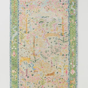 May include: A rectangular rug with a detailed, colorful design. The rug features a light green background with a central scene of trees, flowers, and animals in pink, yellow, and blue. A green border frames the design.