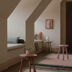 May include: A cozy interior scene featuring a pink rug with a green border, a wooden side table with a pink vase, and a pink stool. A small framed artwork hangs on the wall, and a bench with pillows is visible.
