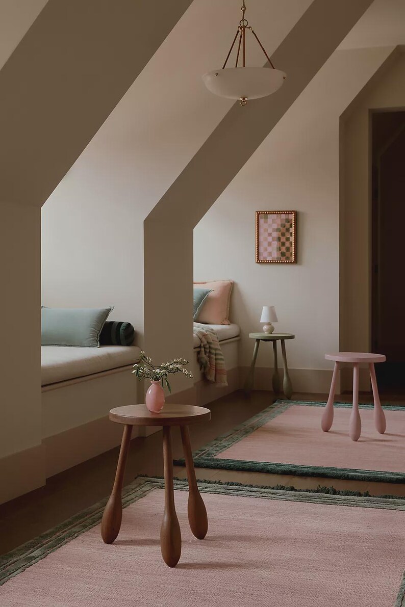 May include: A cozy interior scene featuring a pink rug with a green border, a wooden side table with a pink vase, and a pink stool. A small framed artwork hangs on the wall, and a bench with pillows is visible.