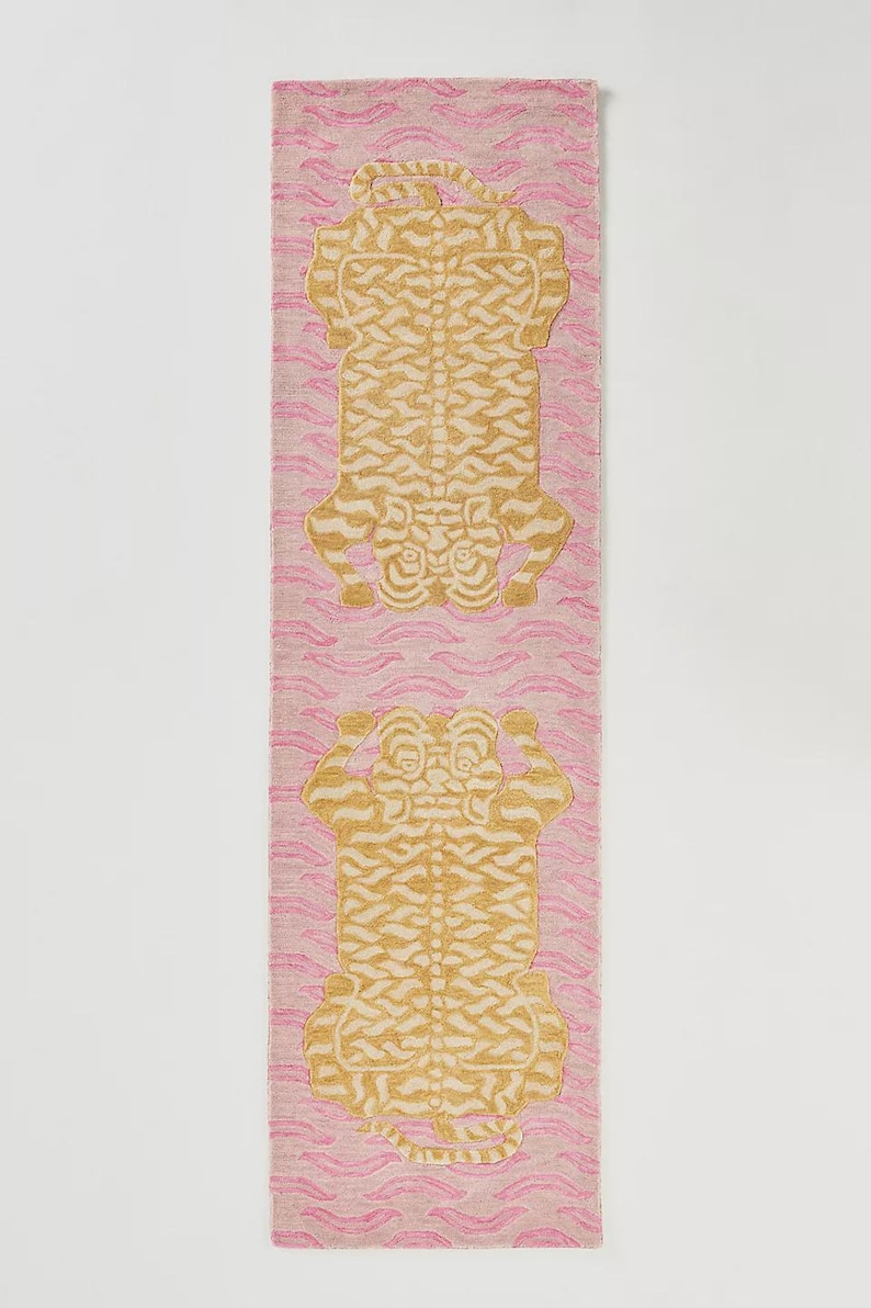 May include: A pink runner rug with a repeating pattern of stylized gold tiger figures. The rug has a wavy pink background. The tigers are facing up and down. The rug is a decorative home accent.