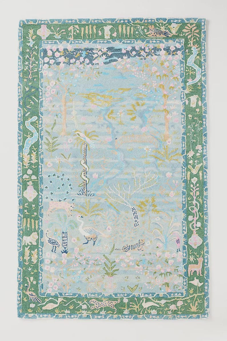 May include: A rectangular rug with a light blue background featuring a whimsical forest scene with trees, birds, and animals. The rug has a green border with decorative patterns. The design includes pink flowers and gold accents.
