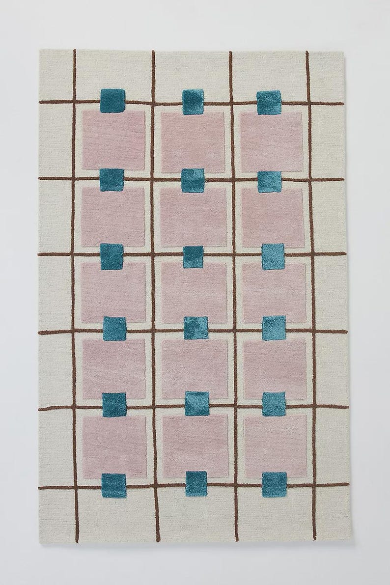 Puede incluir: Alfombra rectangular con fondo crema y un patr&oacute;n de cuadr&iacute;cula de l&iacute;neas marrones. Cuadrados rosas dispuestos en filas y columnas, cada uno con un cuadrado turquesa en el centro. Dise&ntilde;o geom&eacute;trico moderno.