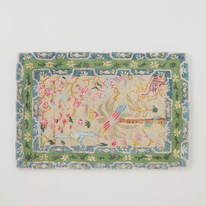 May include: Rectangular rug with a beige background and a floral design. The rug features a green border with a blue trim. The central design includes pink flowers, birds, and other botanical elements. The rug is likely made of a soft, woven material.