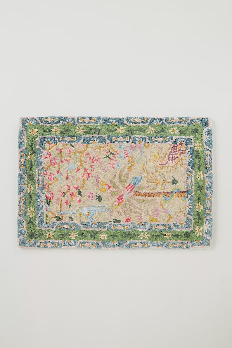 May include: Rectangular rug with a beige background and a floral design. The rug features a green border with a blue trim. The central design includes pink flowers, birds, and other botanical elements. The rug is likely made of a soft, woven material.