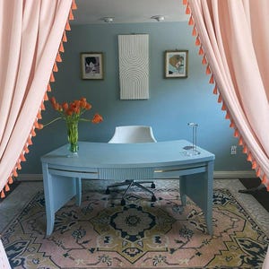 May include: A home office with a light blue desk, a white chair, and a colorful rug. Pink curtains with orange tassels frame the room. Artwork hangs on the blue wall, and a vase of orange flowers sits on the desk.