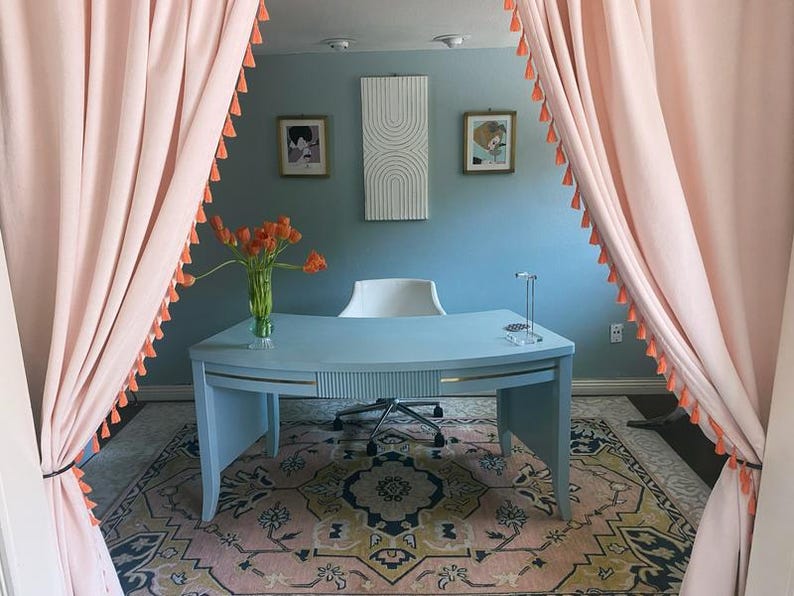 May include: A home office with a light blue desk, a white chair, and a colorful rug. Pink curtains with orange tassels frame the room. Artwork hangs on the blue wall, and a vase of orange flowers sits on the desk.