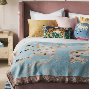 Op de afbeelding: Een bed met een lichtblauwe deken met een paardenmotief, aangevuld met decoratieve kussens. Kussens met bloemen-, tijger- en paardenmotieven. Het bed heeft een mauve hoofdbord en staat op een blauw tapijt met een geblokte rand.