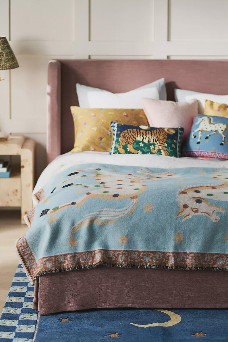 Op de afbeelding: Een bed met een lichtblauwe deken met een paardenmotief, aangevuld met decoratieve kussens. Kussens met bloemen-, tijger- en paardenmotieven. Het bed heeft een mauve hoofdbord en staat op een blauw tapijt met een geblokte rand.