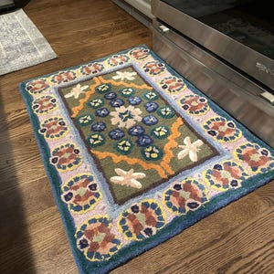 May include: A rectangular, hand-tufted rug with a floral design. The rug features a central field of green with blue and white flower motifs, framed by a border of repeating floral patterns in shades of pink, brown, and blue. The rug's edges are a deep blue.