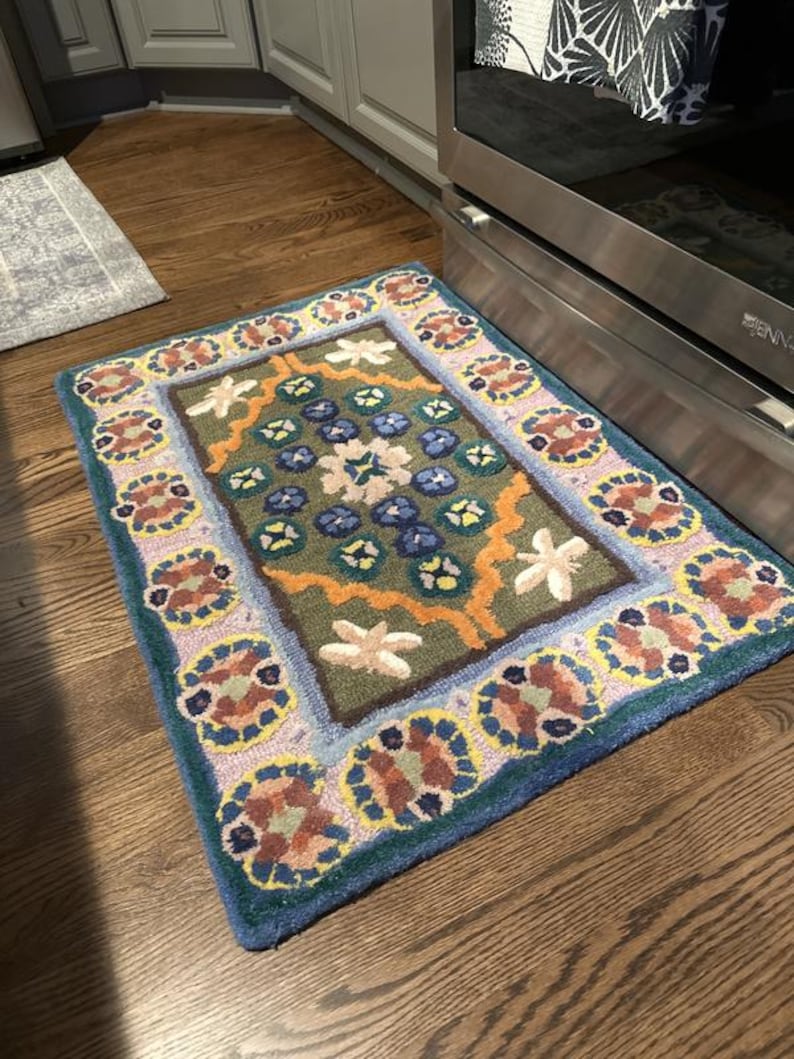 May include: A rectangular, hand-tufted rug with a floral design. The rug features a central field of green with blue and white flower motifs, framed by a border of repeating floral patterns in shades of pink, brown, and blue. The rug's edges are a deep blue.