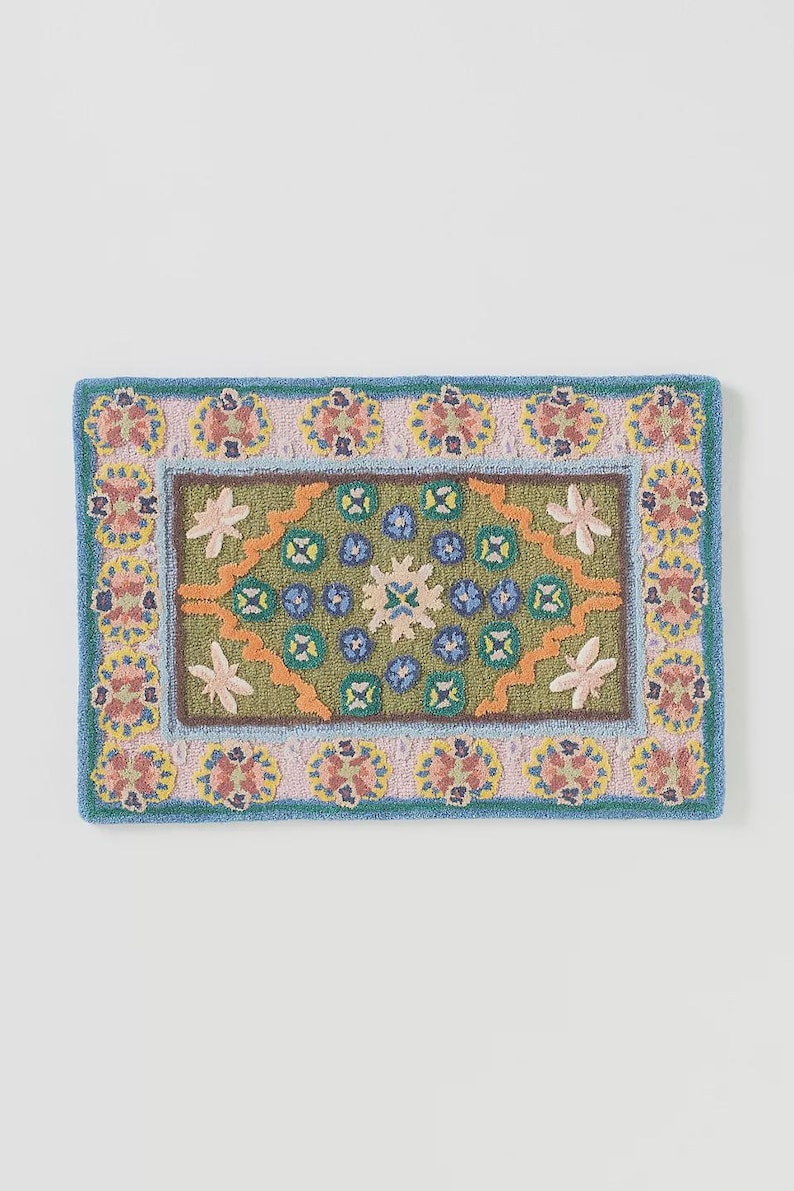 May include: Rectangular, hand-tufted rug with a floral design. The rug features a green center with blue, orange, and white floral accents. The border is light pink with yellow and red floral patterns, framed by a blue edge.