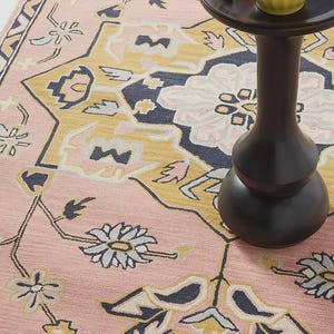 May include: A pink area rug with a floral pattern in shades of yellow, navy, and cream. A small black side table holds a bowl of popcorn and a glass of pink liquid. The rug's design features stylized flowers and geometric shapes.