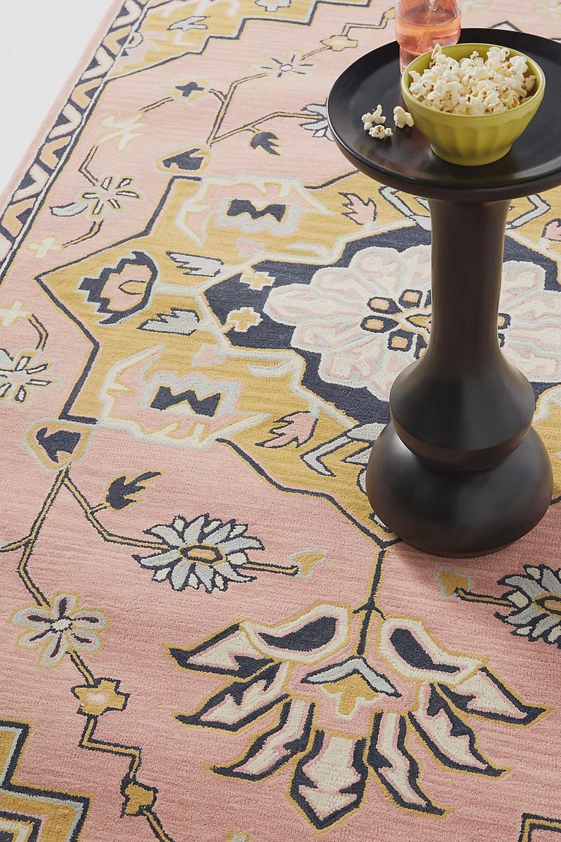 May include: A pink area rug with a floral pattern in shades of yellow, navy, and cream. A small black side table holds a bowl of popcorn and a glass of pink liquid. The rug's design features stylized flowers and geometric shapes.